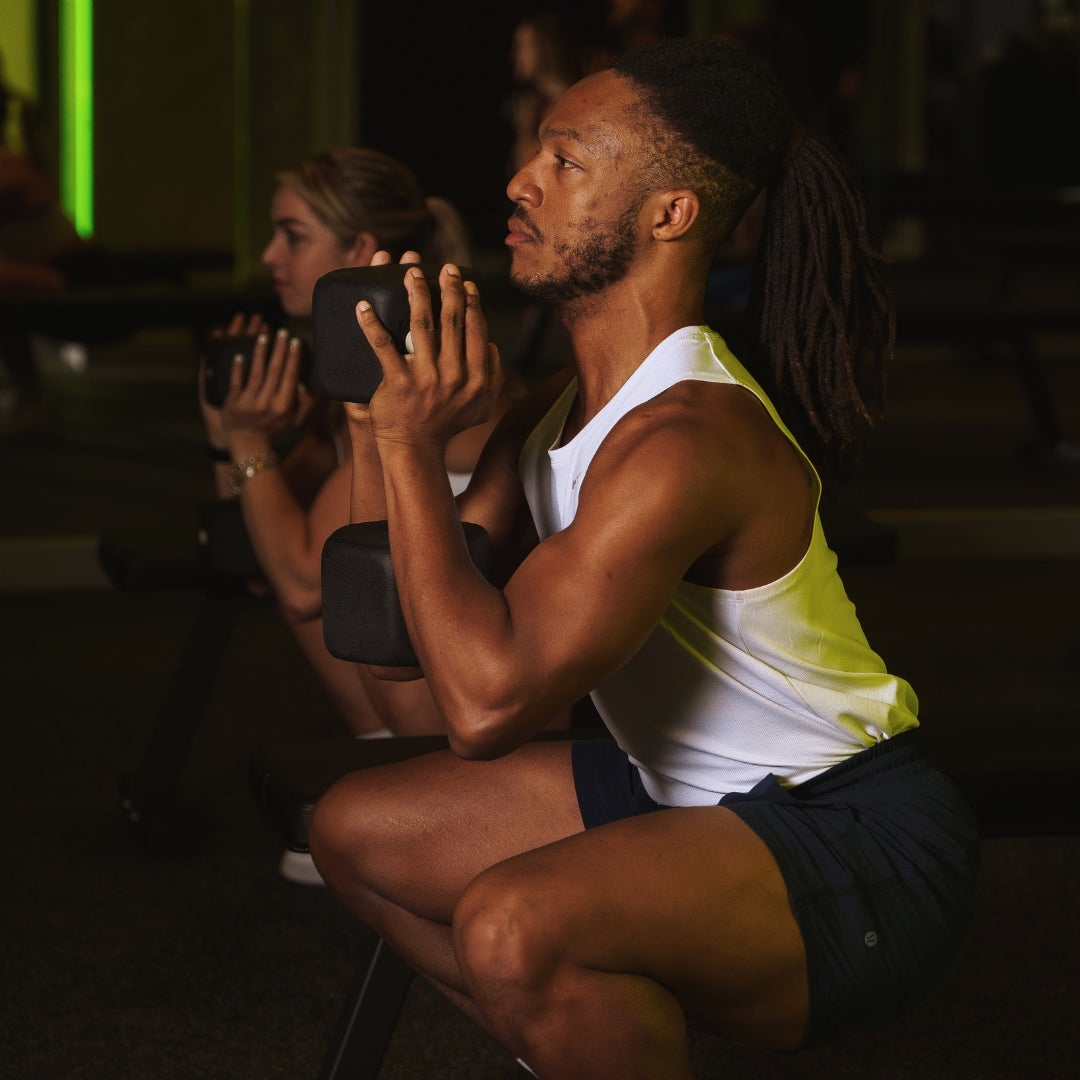 Man in a white tank top and black shorts squatting in a Strength studio while holding a dumbbell